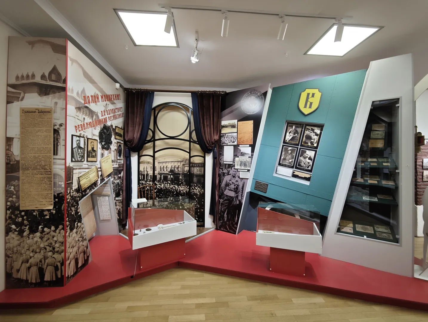 The exhibition area is predominantly red, with the walls covered in historical photos and slogans. Display cases contain various documents and memorabilia, and the background wall features large historical scene paintings. The layout is compact, with abundant information.