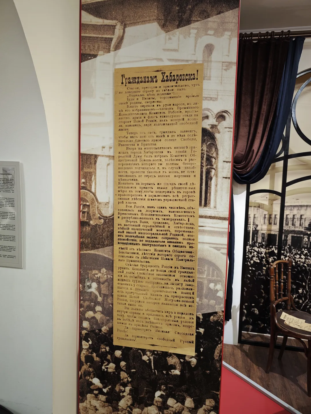 An old newspaper is displayed on the wall, with Russian text and black-and-white photographs. Next to the newspaper is an image of a building, set against a light-colored wall. To the right, there are dark curtains and a metal-framed window.