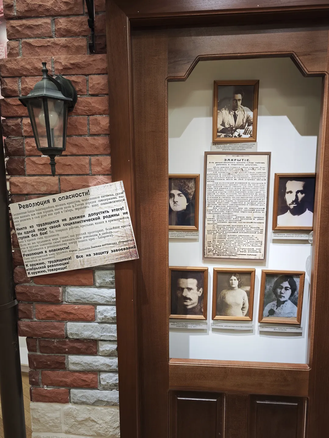 Several black-and-white photos and a notice are displayed inside a wooden door next to a brick wall. The people in the photos have serious expressions, and the notice pertains to revolutionary themes.