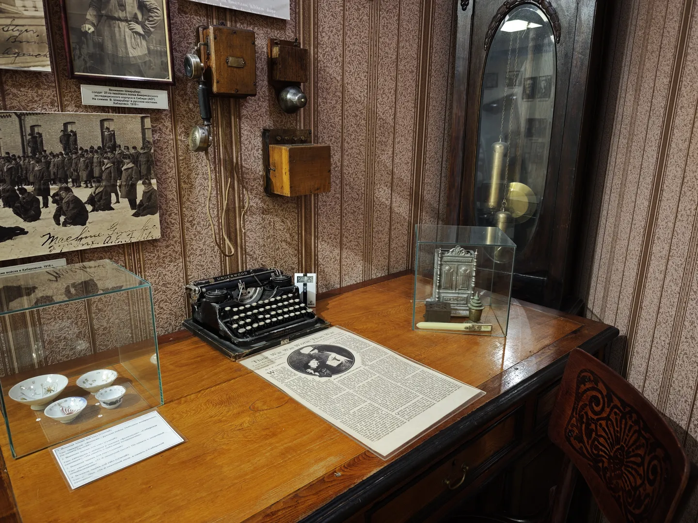 A wooden table is set with an old-fashioned typewriter and a newspaper. Several black-and-white photos are hung on the wall, and a telephone is placed nearby.