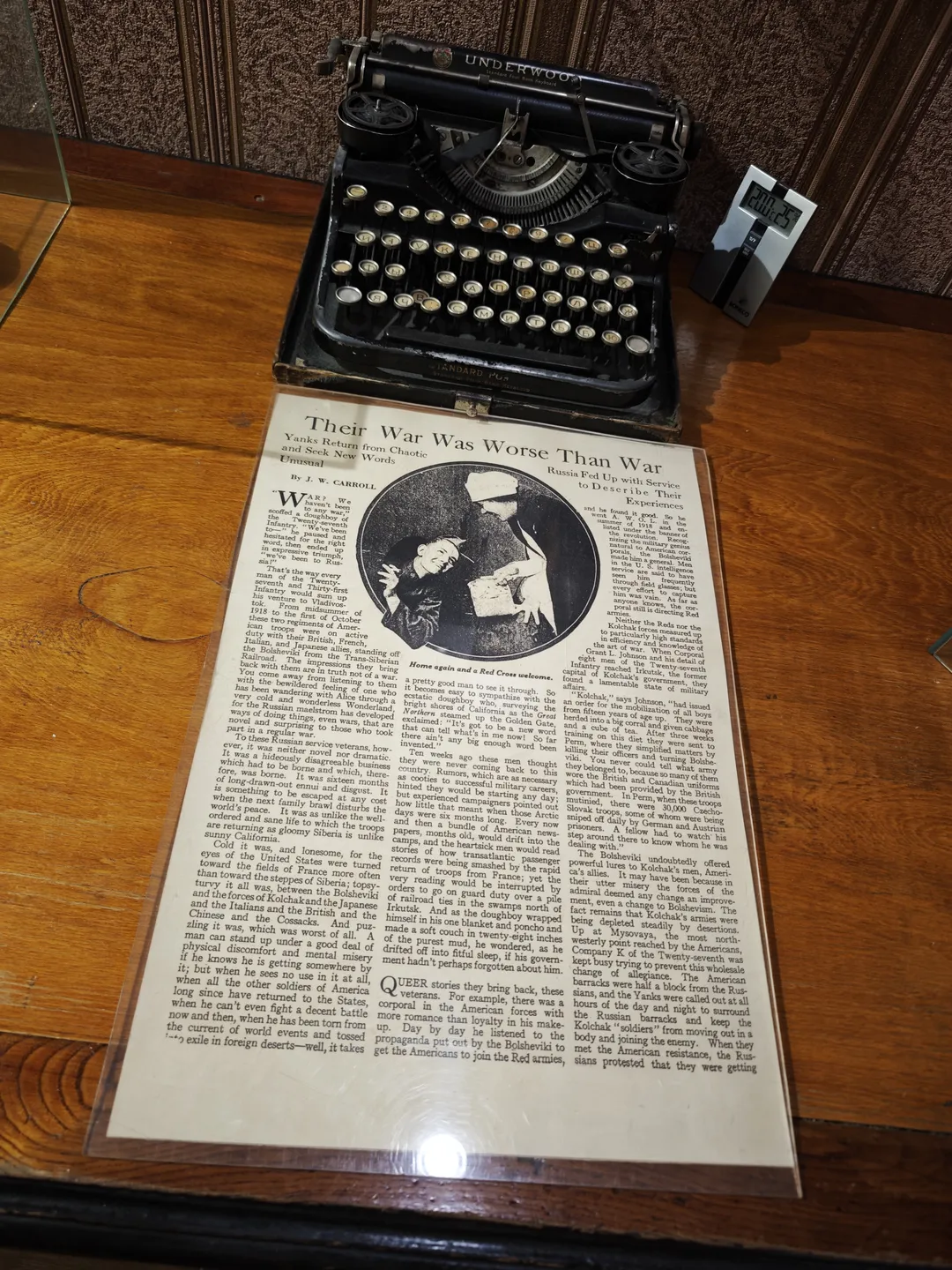 A newspaper titled “Their War Was Worse Than War” is placed on a wooden table. Next to it is a black typewriter, set against a wallpapered background.