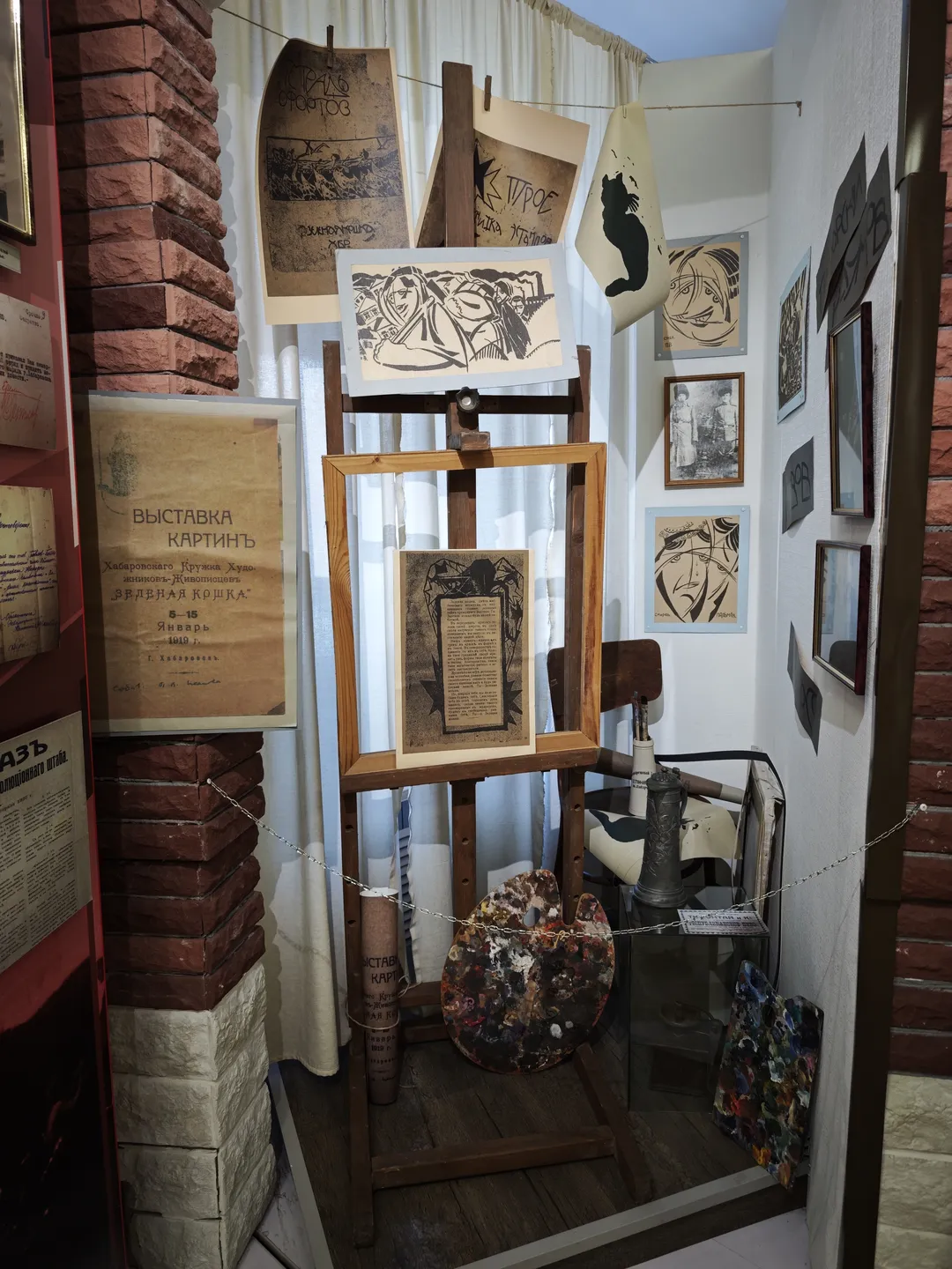 The exhibition area displays paintings and related items. Several paintings are hung on the wall, and in front of them are easels and palettes.