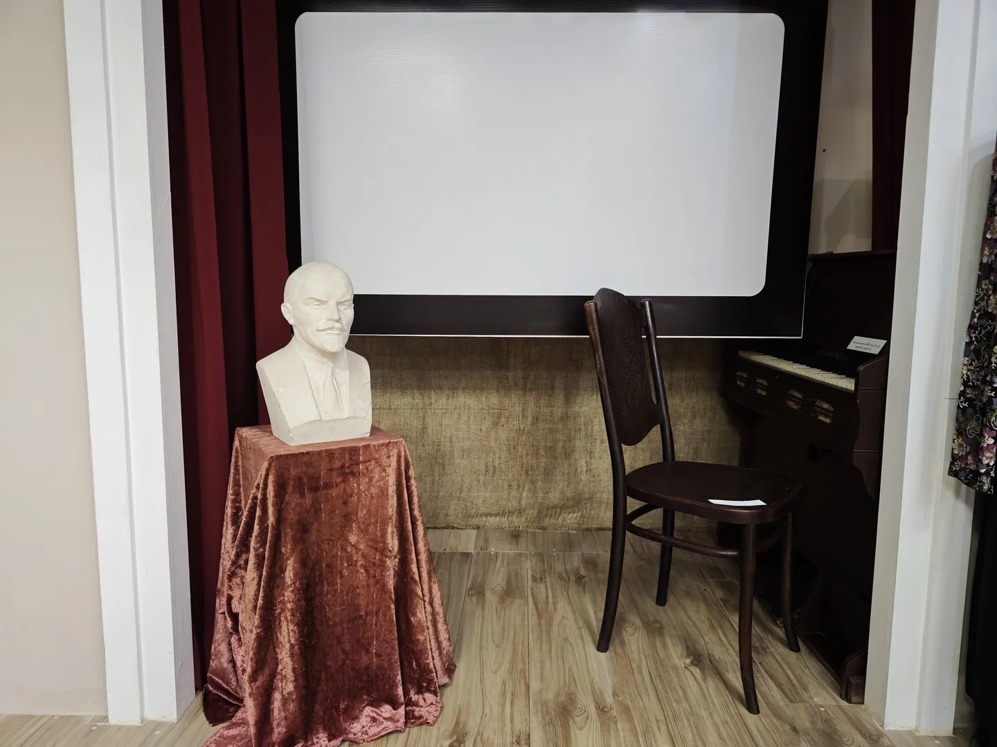 A white statue is placed on a table draped with red fabric, accompanied by a chair and a piano. The background wall features a blank projection screen.