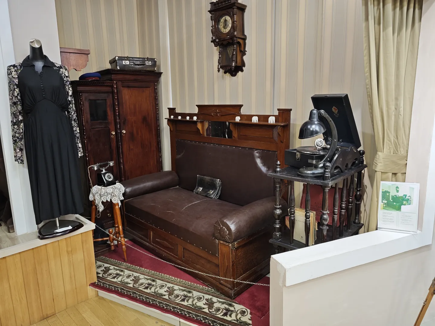 A brown leather sofa is placed in the room, with a small table next to it that holds an old typewriter. On the wall is a clock, and nearby stands a wooden wardrobe and a telephone model.