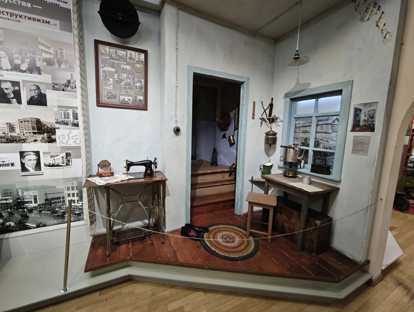 A small indoor scene model displays an entryway. On the left is a table with a sewing machine, and on the right is another small table with a teapot and cups, next to a window. A round rug is laid on the floor.
