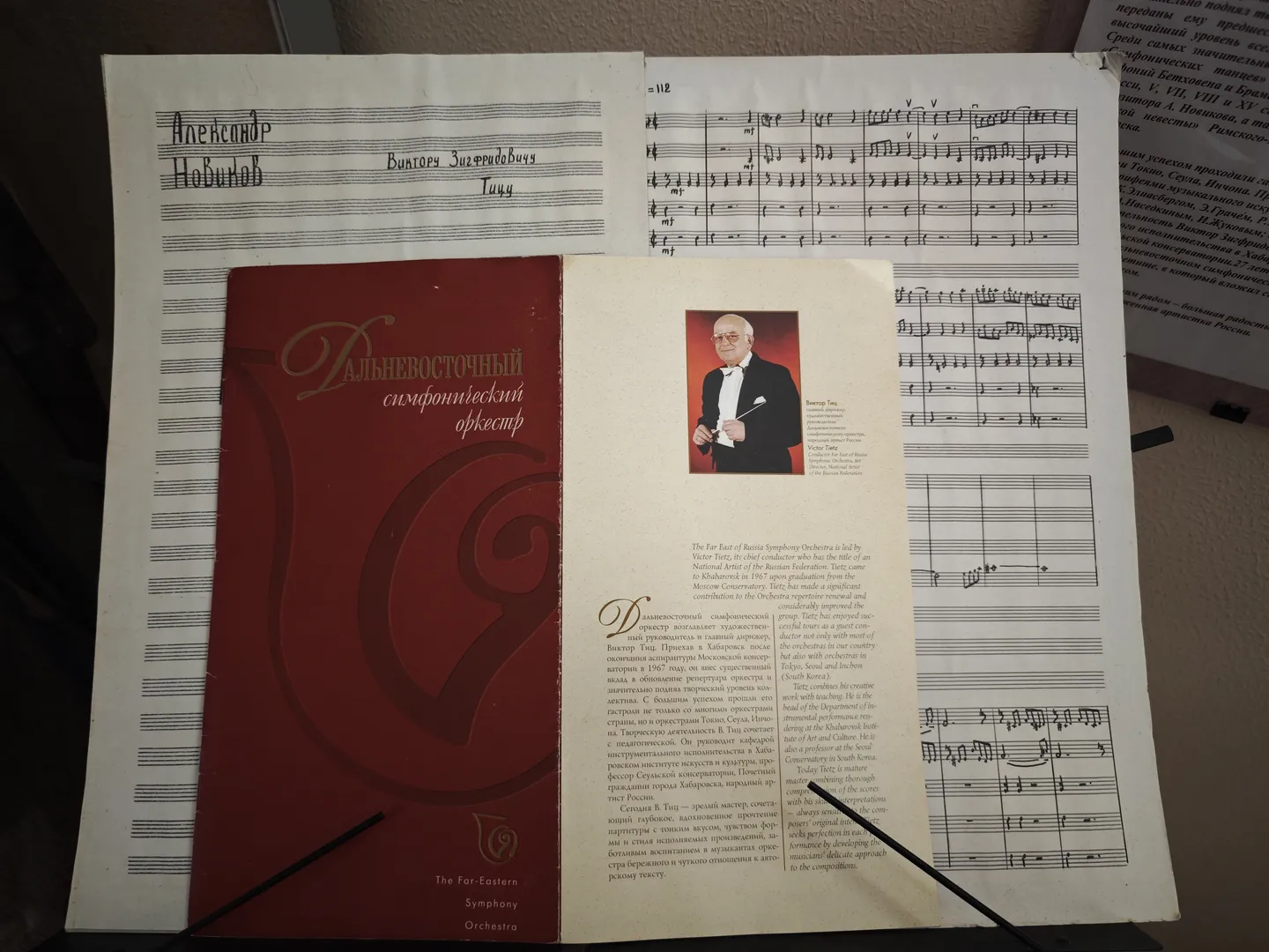 A red-covered book is placed on a music stand, with a photo of a man in a suit on the cover. The pages of the music score are opened, displaying musical notation and notes. Russian text is visible above the music stand.