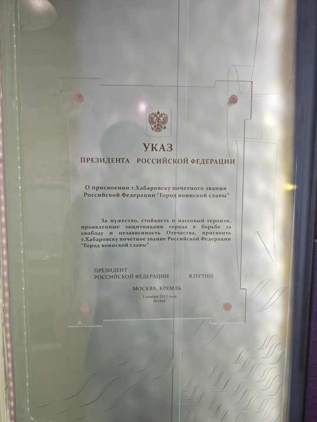 A document is displayed inside a transparent glass case, with the national emblem and the word “УКАЗ” at the top. The document mentions the granting of the title “City of Military Glory” to Khabarovsk. Below the document is the president’s signature and the date.