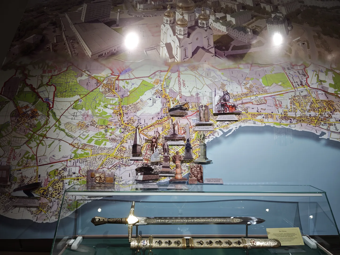 A city map model is displayed inside a glass case, featuring landmarks and streets. In front of the map, several decorative swords and information plaques are displayed. The center of the map features a large church model.