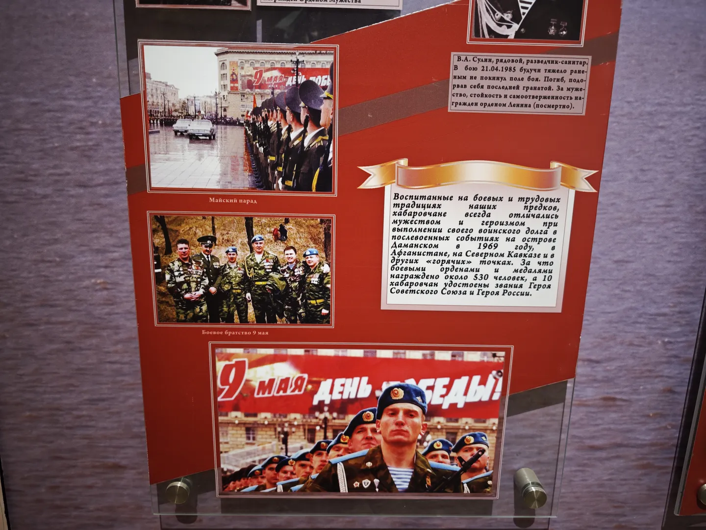 An exhibition board displays photos of soldiers, including group marches and collective portraits. The board’s text describes the soldiers’ heroic deeds and honors. At the bottom of the board is a photo from a “Victory Day” celebration.