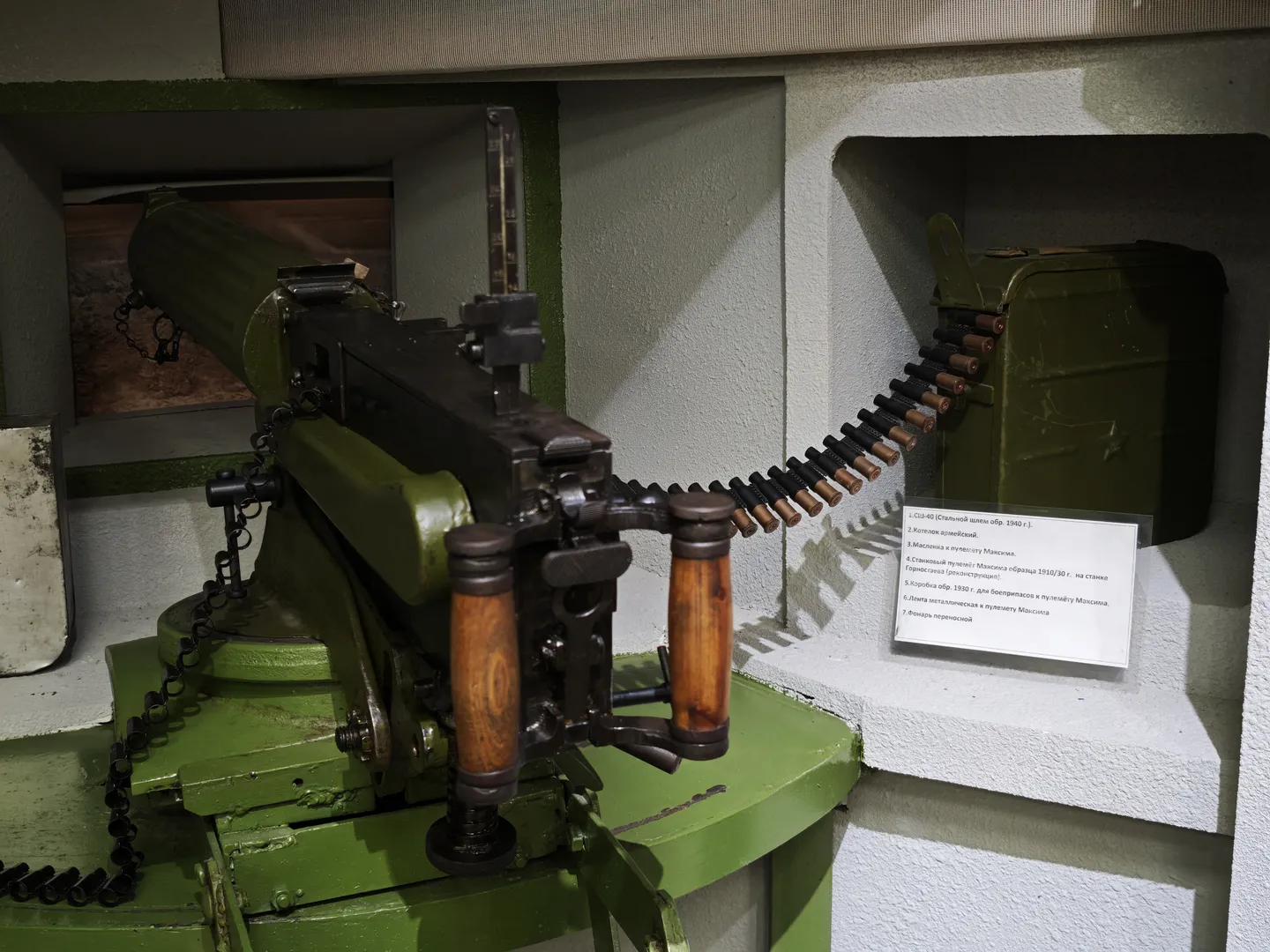 A green machine gun is displayed inside a glass case, next to a belt of ammunition and an ammo box. The machine gun has a complex design, featuring a wooden grip and aiming device. A white label in front of the display provides explanatory text.