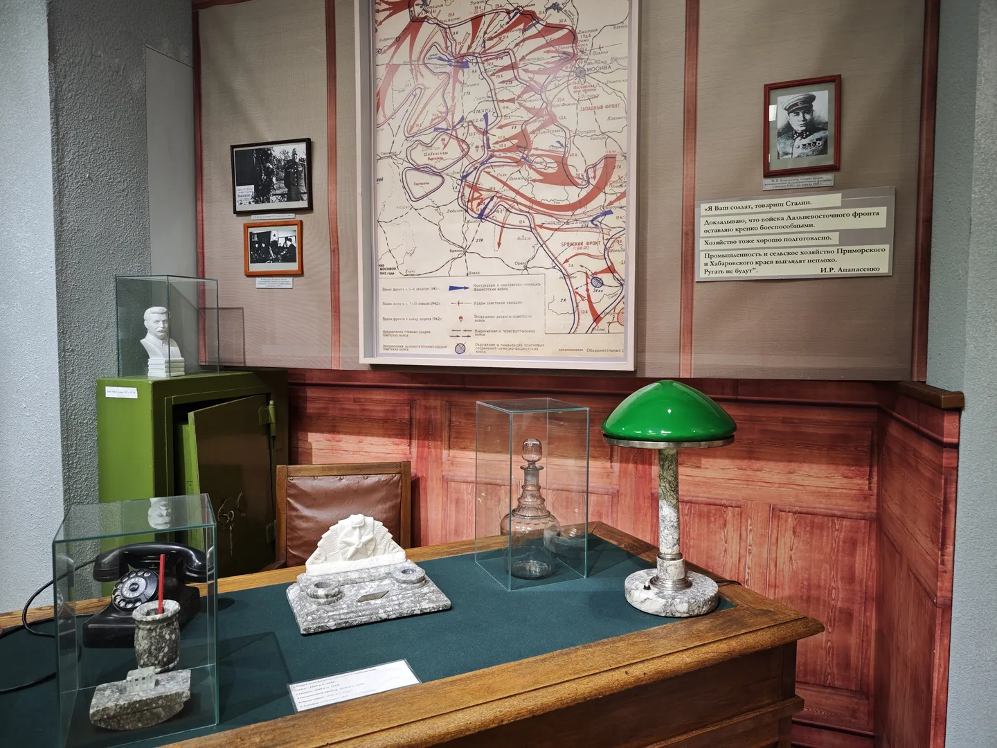 A military map is hung on the wall, with a wooden table beneath it. The table is equipped with a telephone, desk lamp, and other items. Several glass display boxes containing small objects are also on the table. The map uses different colors to mark strategic positions.