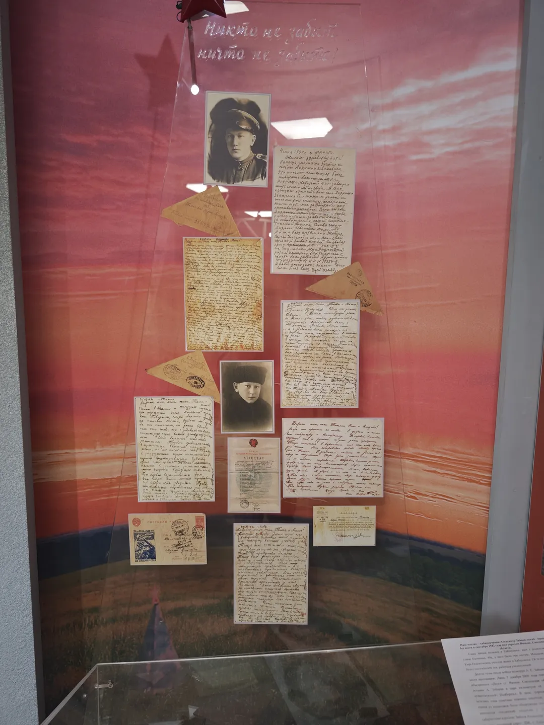 The display case contains several handwritten letters and photographs, set against a red gradient background. The letters and photos are neatly arranged, with two black-and-white portraits hanging above. A white label at the bottom provides relevant information.