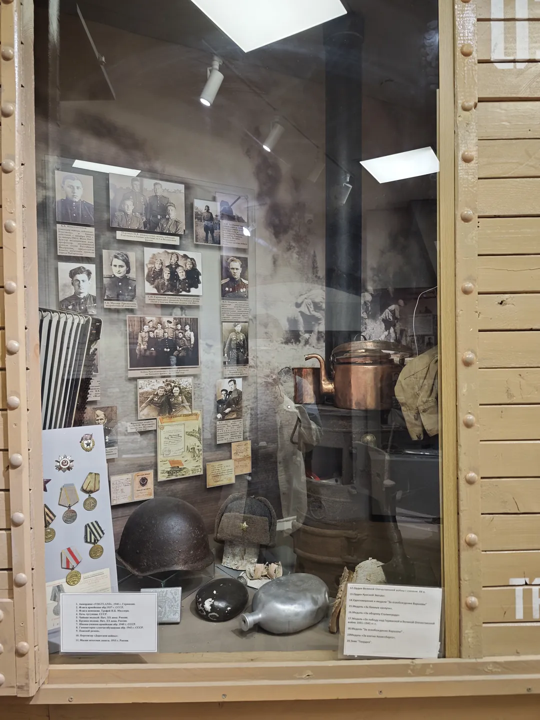 The display case contains military items such as helmets, caps, and water bottles. The background wall is adorned with black-and-white photos and documents. On the left side of the case, several medals and honors are displayed, while the right side features detailed text descriptions. The case is designed simply to highlight the historical artifacts.