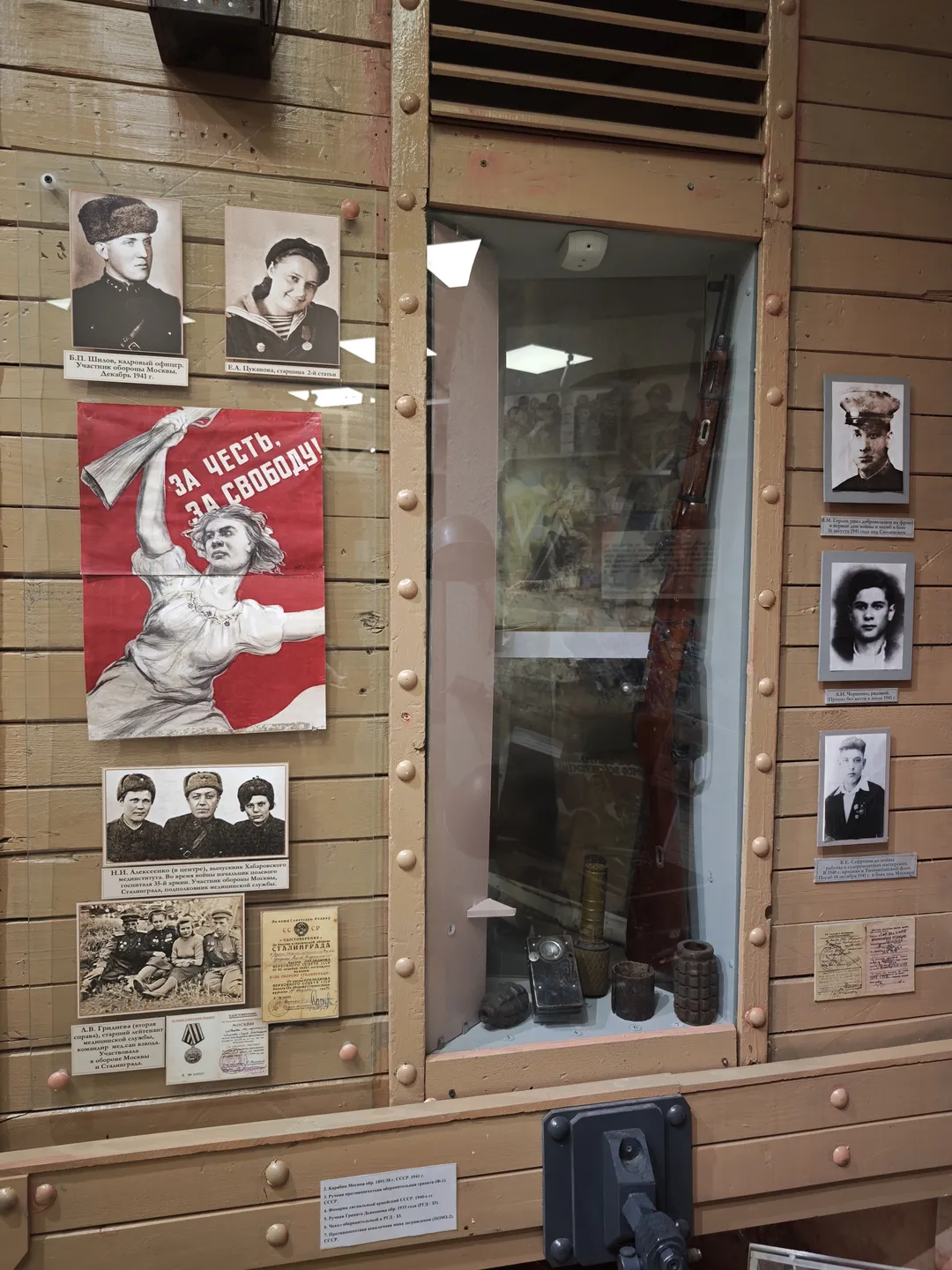 Inside a wooden display case, there is a rifle and several grenade models. Black-and-white portraits of people are attached to the sides of the case. Above the case, a red propaganda poster in Russian hangs. The case has a vintage design with rich details.