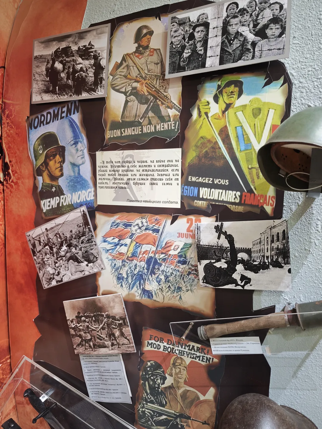 The wall displays several World War II propaganda posters and photographs, depicting soldiers and war scenes. The posters are colorful, and the text is in multiple languages. In the corner of the wall, a metallic object, possibly a weapon part, is placed.