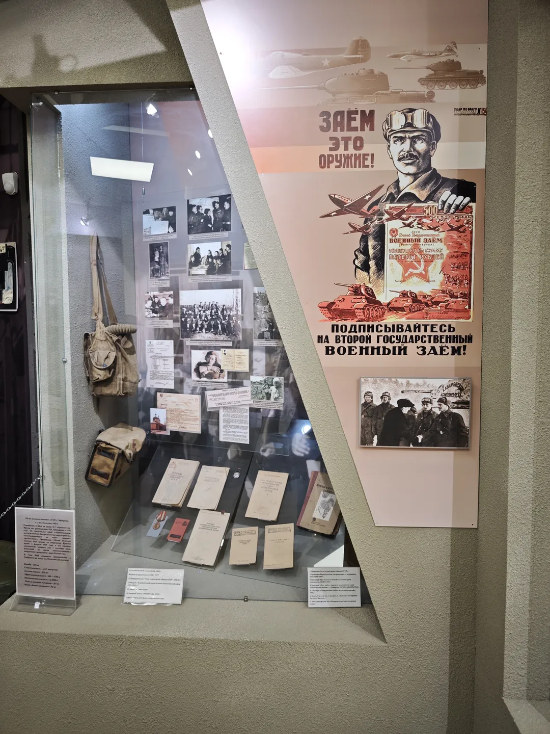 The display case showcases several black-and-white photos and documents. Above the case, a large propaganda poster features a helmeted figure with tanks and airplanes in the background. The case has a modern design with substantial information.