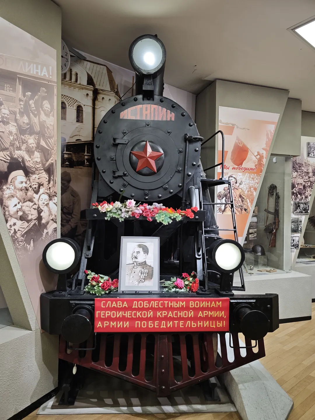 A black steam train model is placed at the center of the exhibition hall. The train’s front is decorated with a red star and floral patterns. A portrait of a person hangs above the train, with a red slogan beneath it. The background wall features several historical photos.