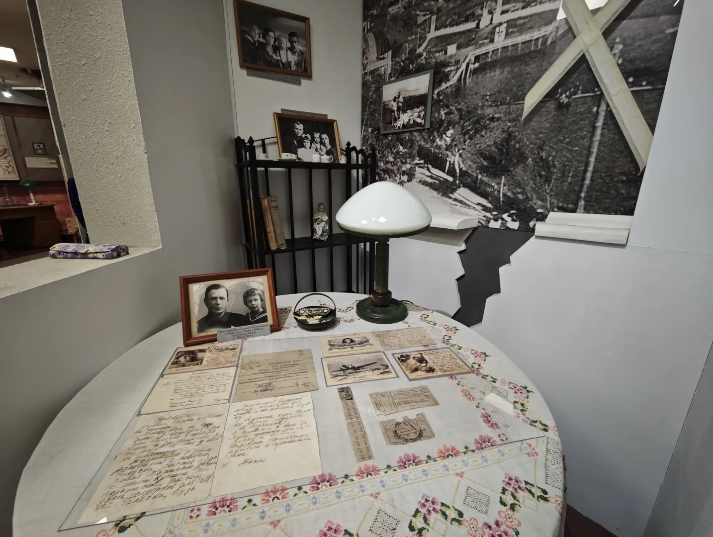A round table with a patterned cloth holds several letters and photos. The table also has a desk lamp and a small basket. On the wall behind, several family portraits are displayed. The overall scene is cozy and rich in historical ambiance.