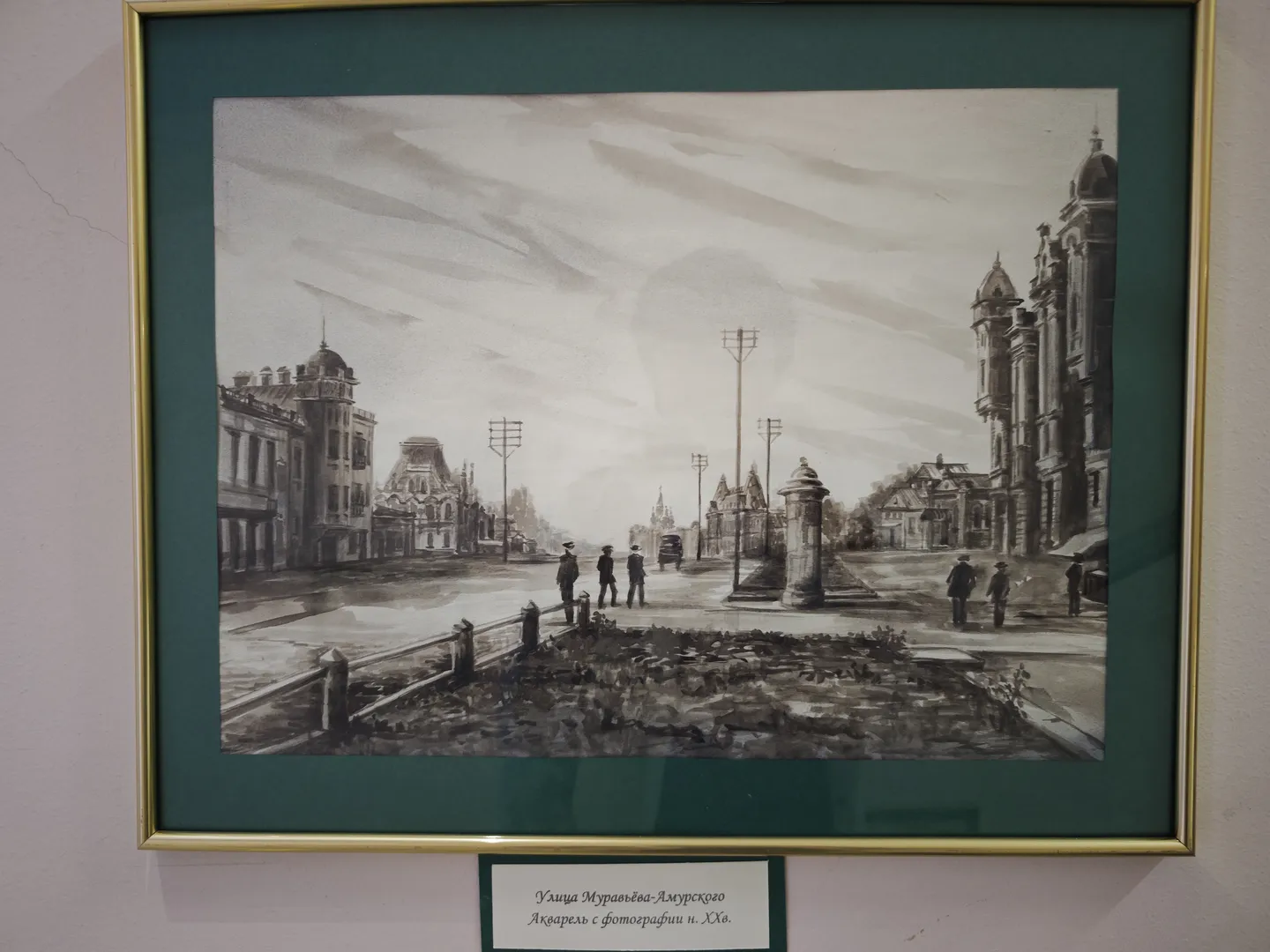 A black-and-white cityscape painting is hung on the wall, framed in gold. The painting depicts streets, buildings, and pedestrians, with clouds flowing in the sky. Below the painting is a white label providing information about the artwork.