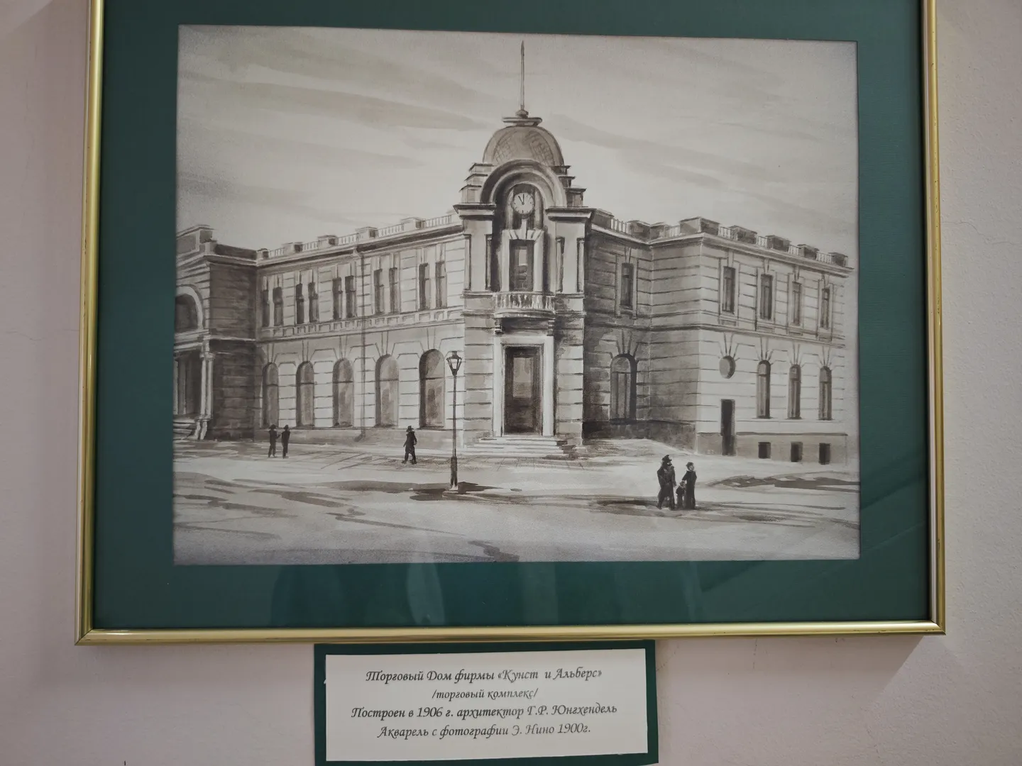 A black-and-white architectural painting depicting a large multi-story building with several people standing in front. The painting is framed in gold, with Russian explanatory text below.