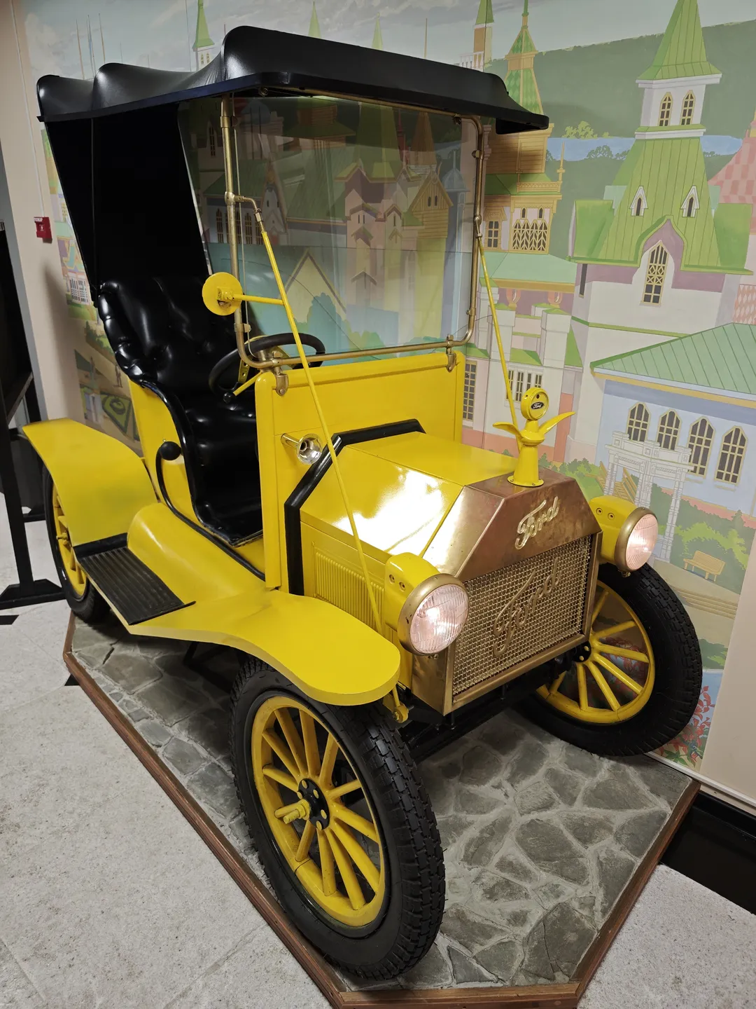 A yellow vintage car model, with a yellow body, black seats, and a steering wheel. The background features a city mural.