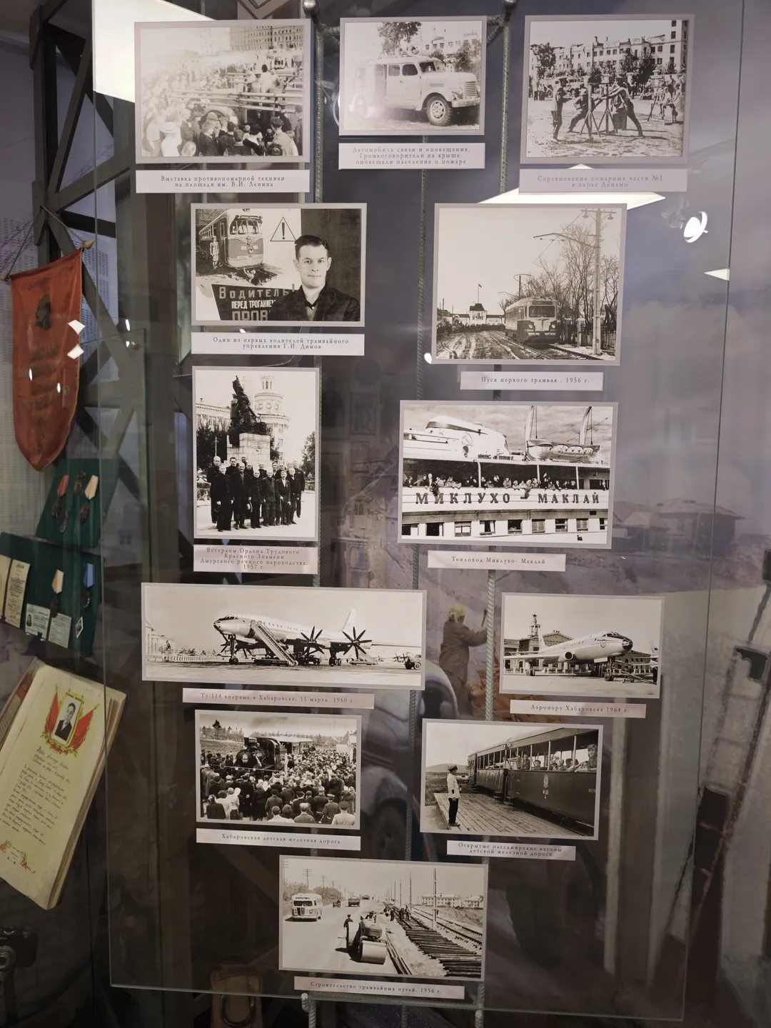 A glass display case containing several black-and-white photos showcasing various historical scenes and figures. A red flag is hung on the left side, with documents and books placed on the right.
