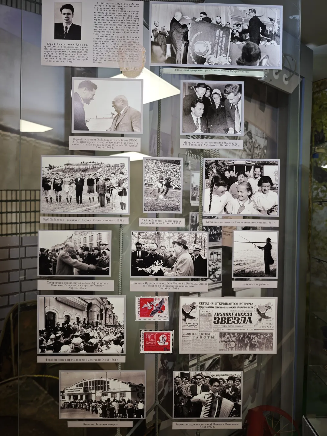 Another glass display case with several black-and-white photos, including group portraits, sports activities, and daily life scenes. Below the case are stamps and newspaper clippings.