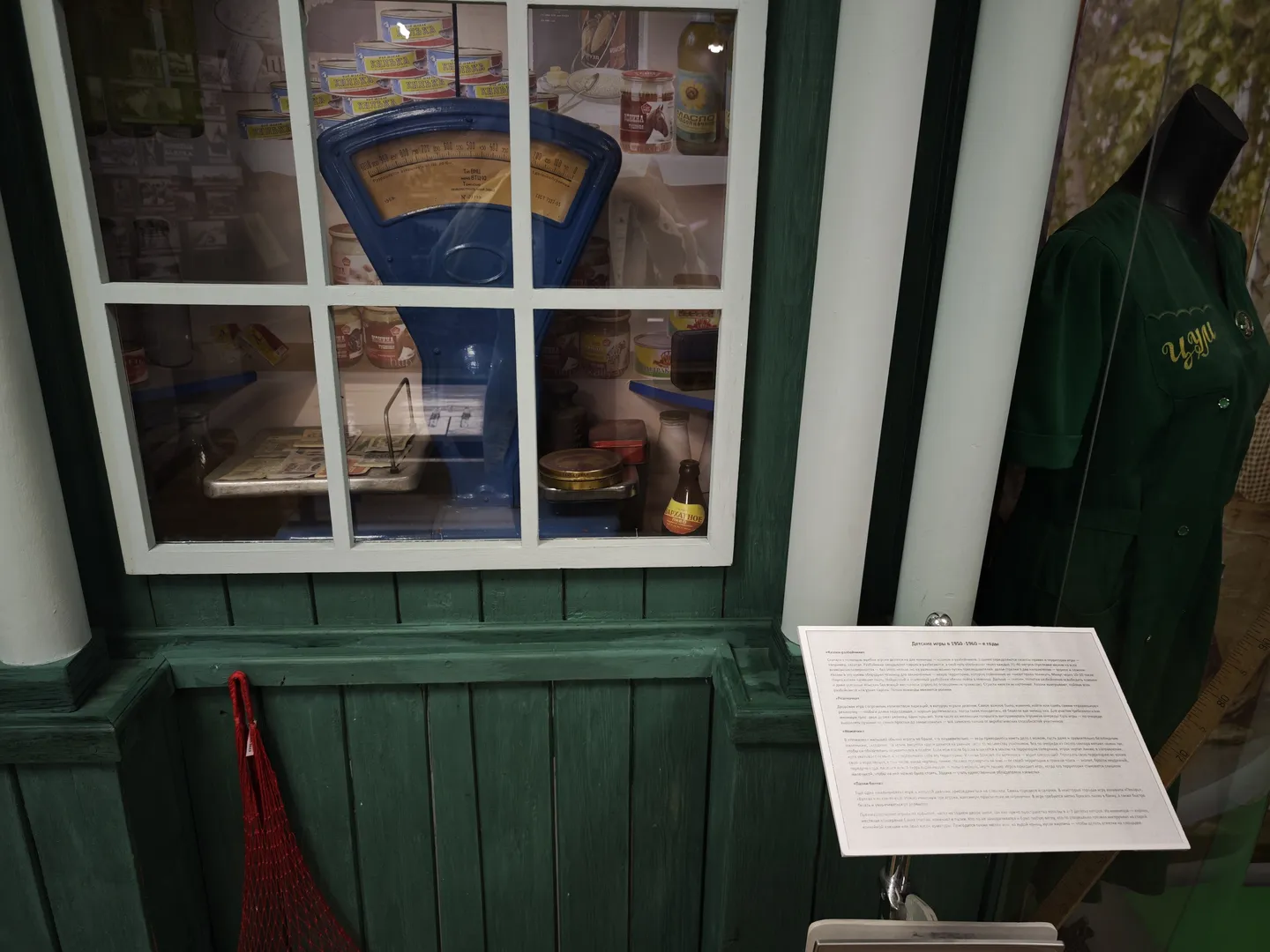 A green wooden counter with a blue scale and canned foods on top. Beside the counter is a mannequin dressed in a green uniform.