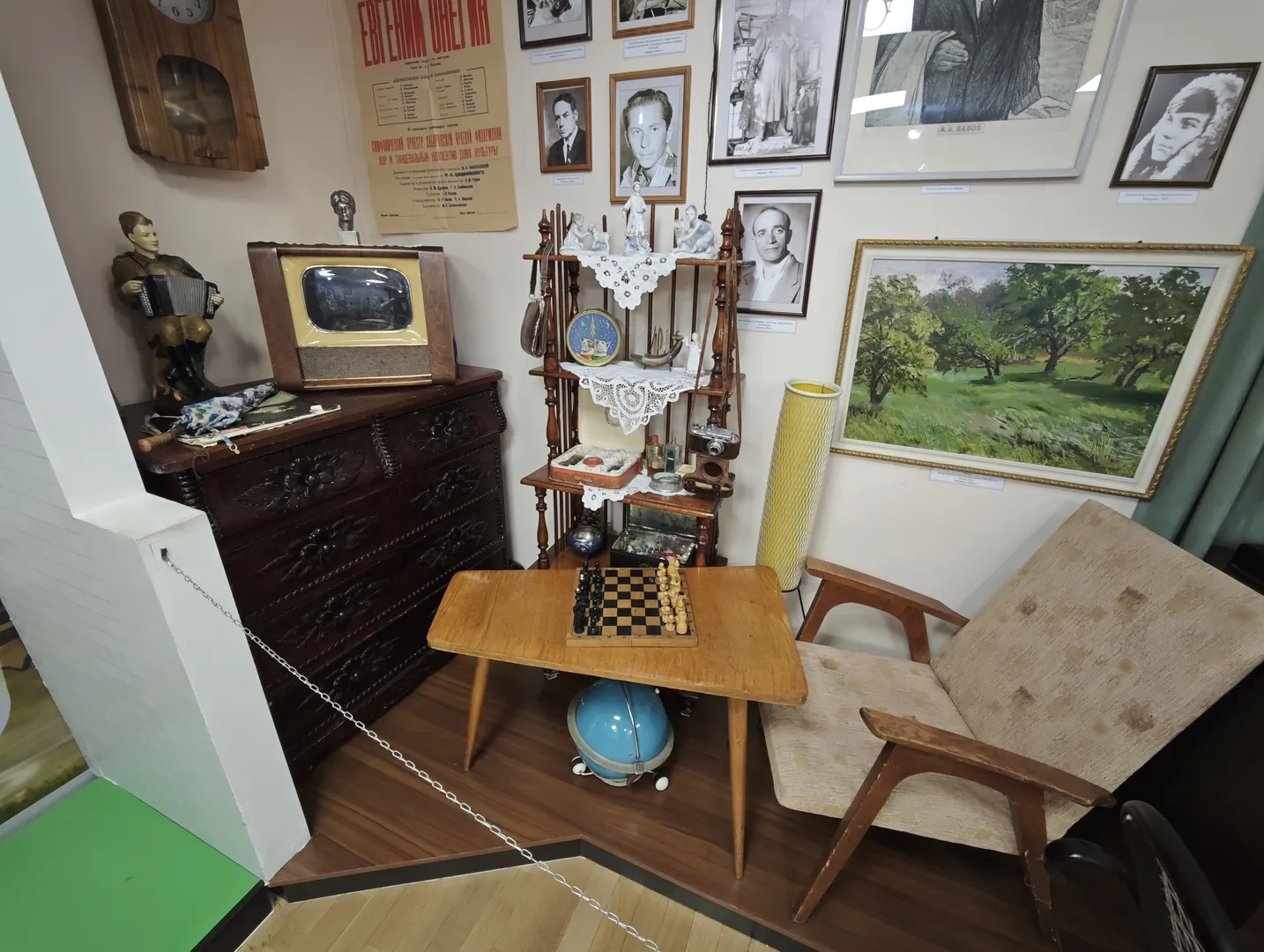 An indoor scene with a wooden table, a chair, and an old-fashioned television. The wall is adorned with several black-and-white photos and a landscape painting.
