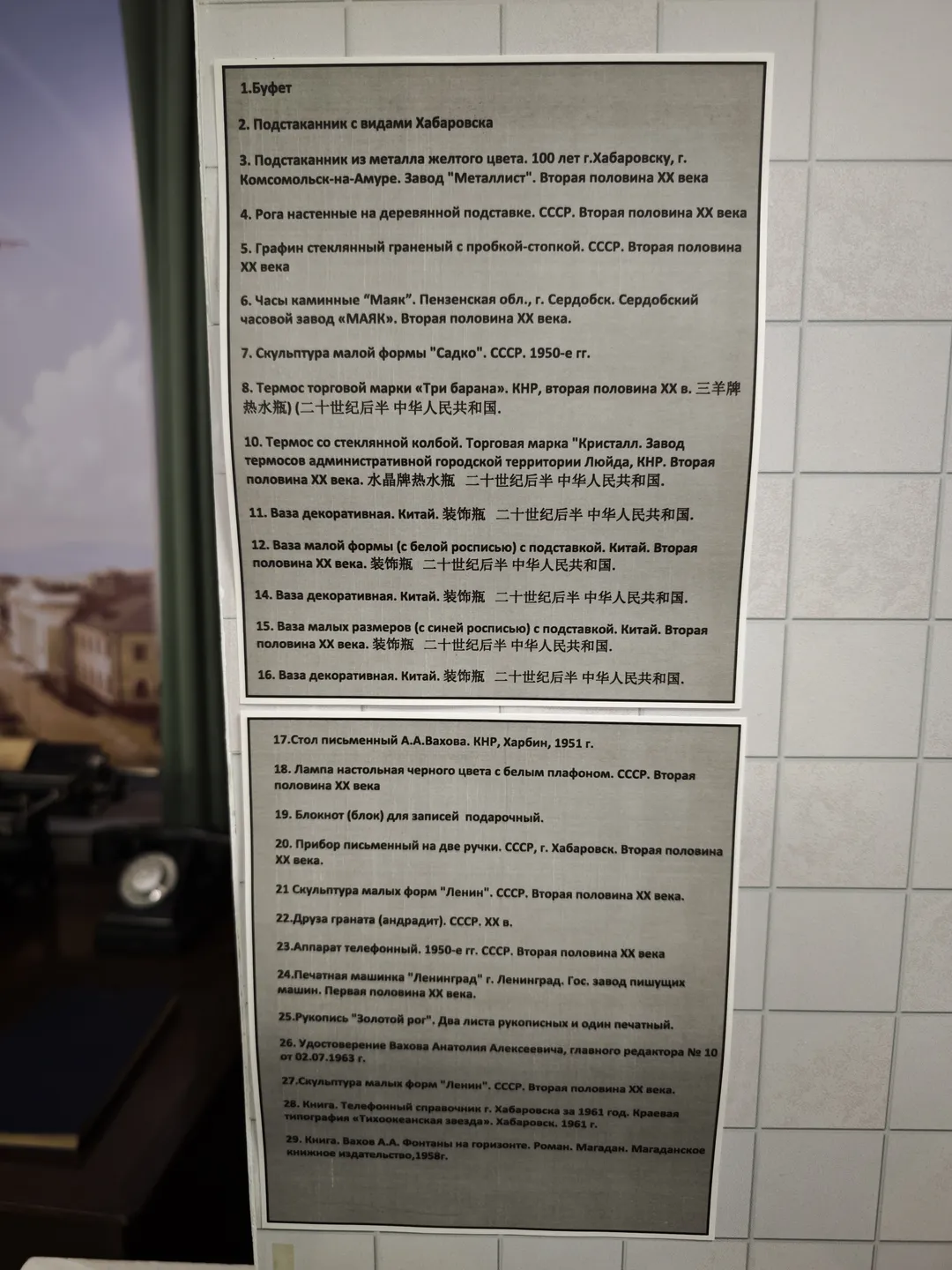 Two information boards with Russian text listing the names and descriptions of various items. The background is a light-colored tiled wall.