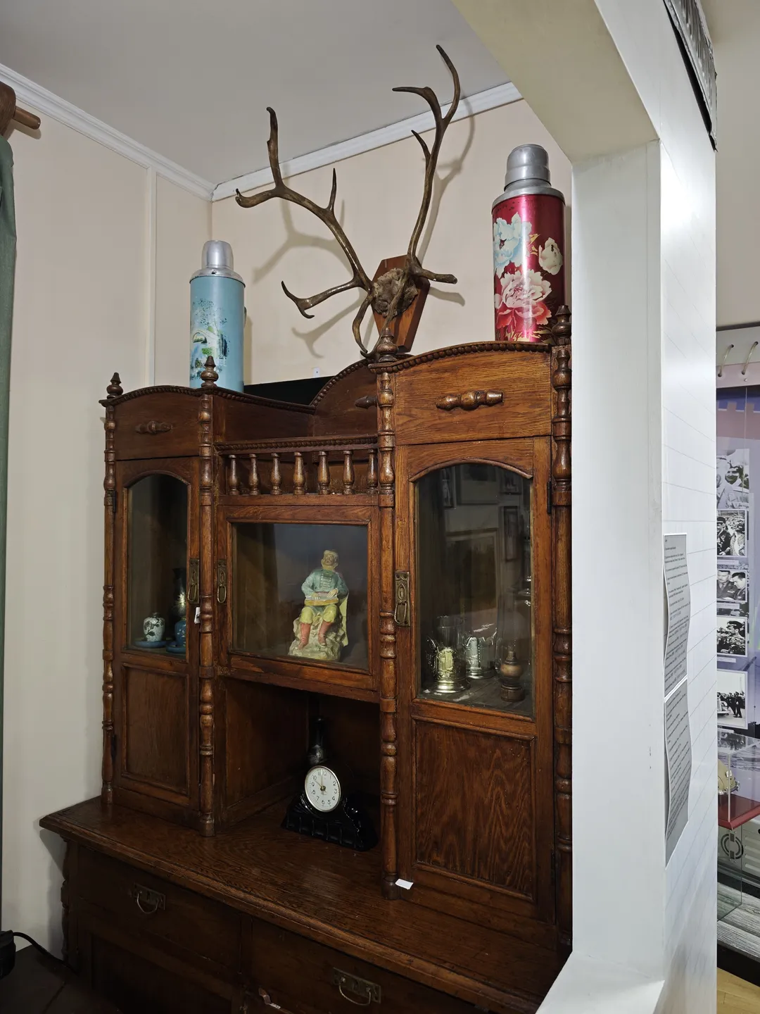 A wooden display cabinet with glass doors containing various decorative items and utensils. Two thermos bottles and a pair of antlers are placed on top of the cabinet.