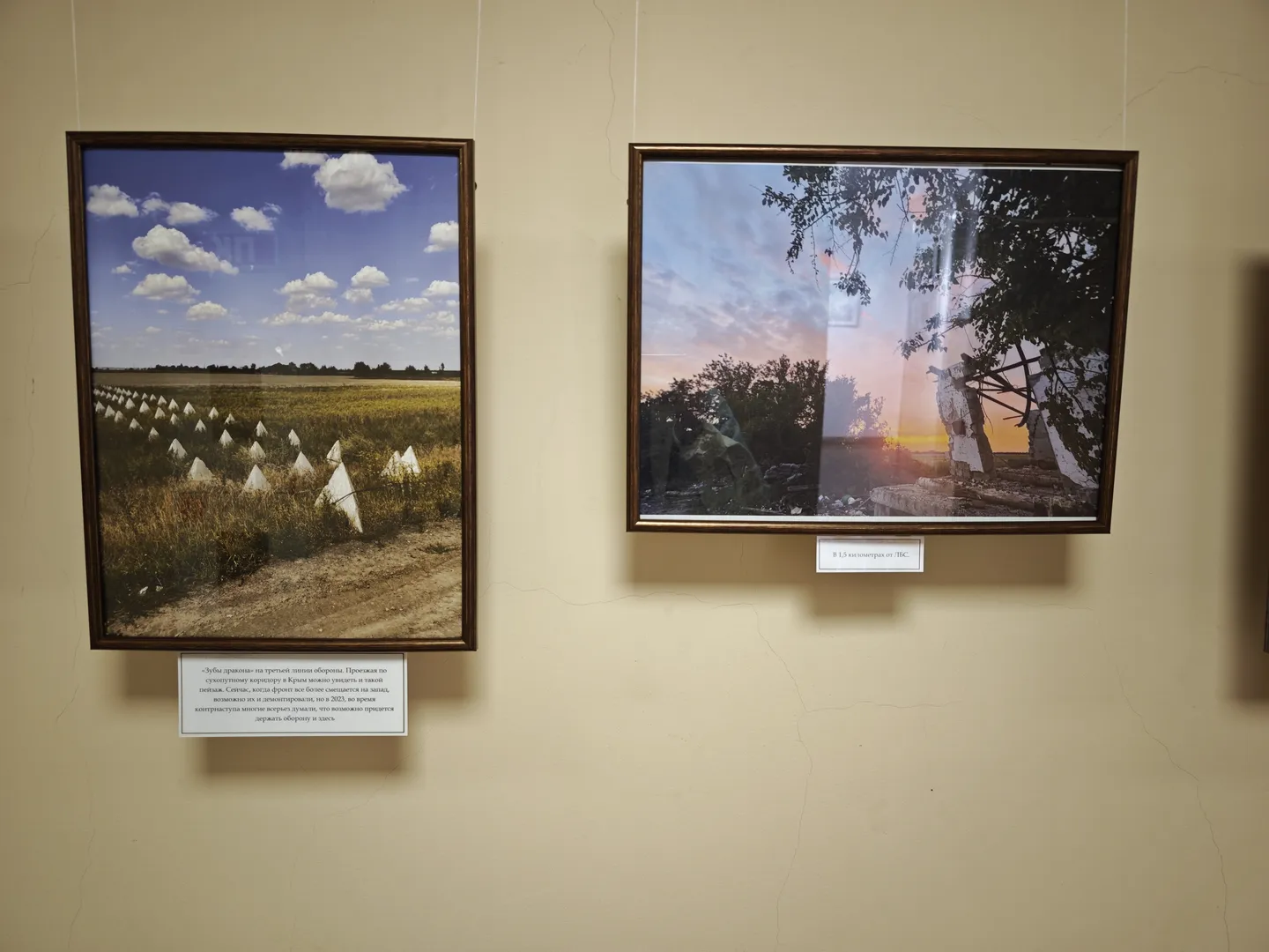 Two paintings hung on the wall. On the left, a field with neatly arranged white conical objects; on the right, trees and abandoned structures at sunset. Each painting has a white label with a description below.
