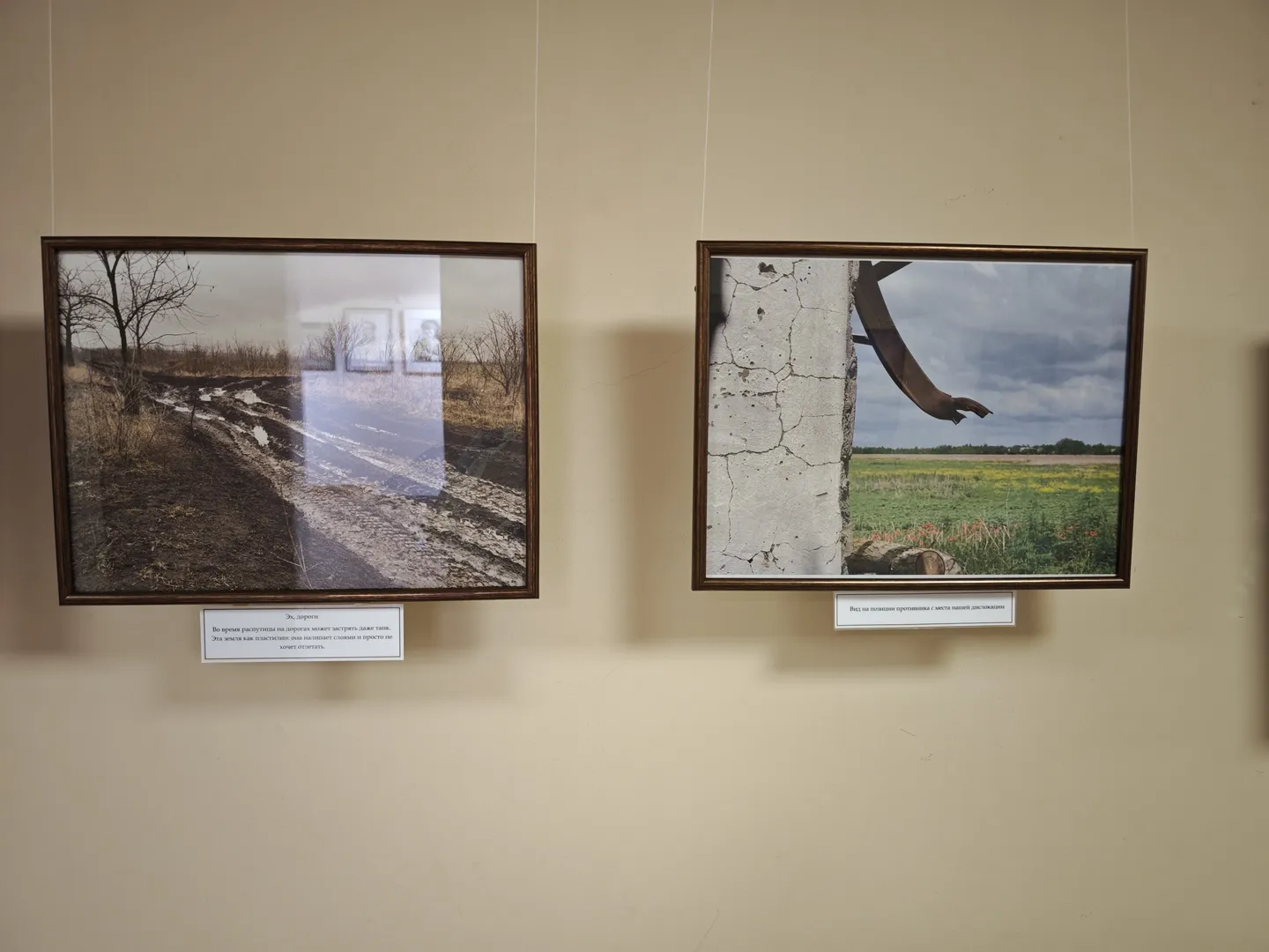 Two paintings on the wall. On the left, a muddy road with bare trees; on the right, a cracked wall and distant fields. Each painting has a white label with a description below.
