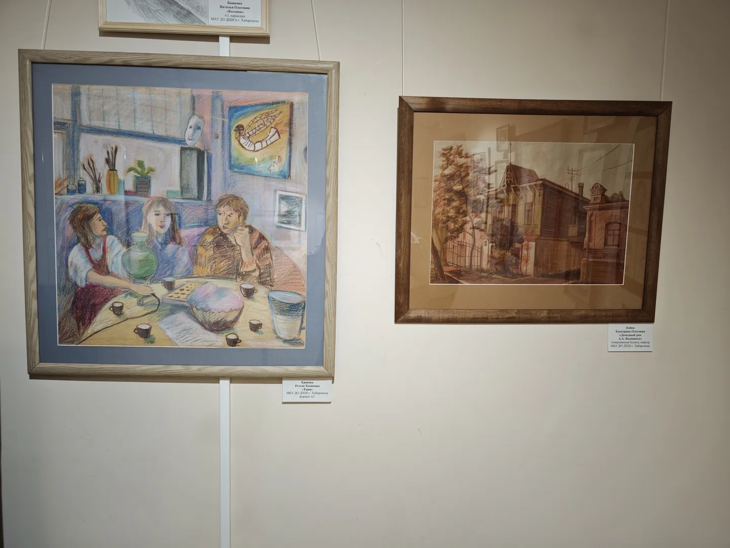 Two paintings hung on the wall. On the left, three people in an indoor scene; on the right, a street scene of an old building. Each painting has a white label with a description below.