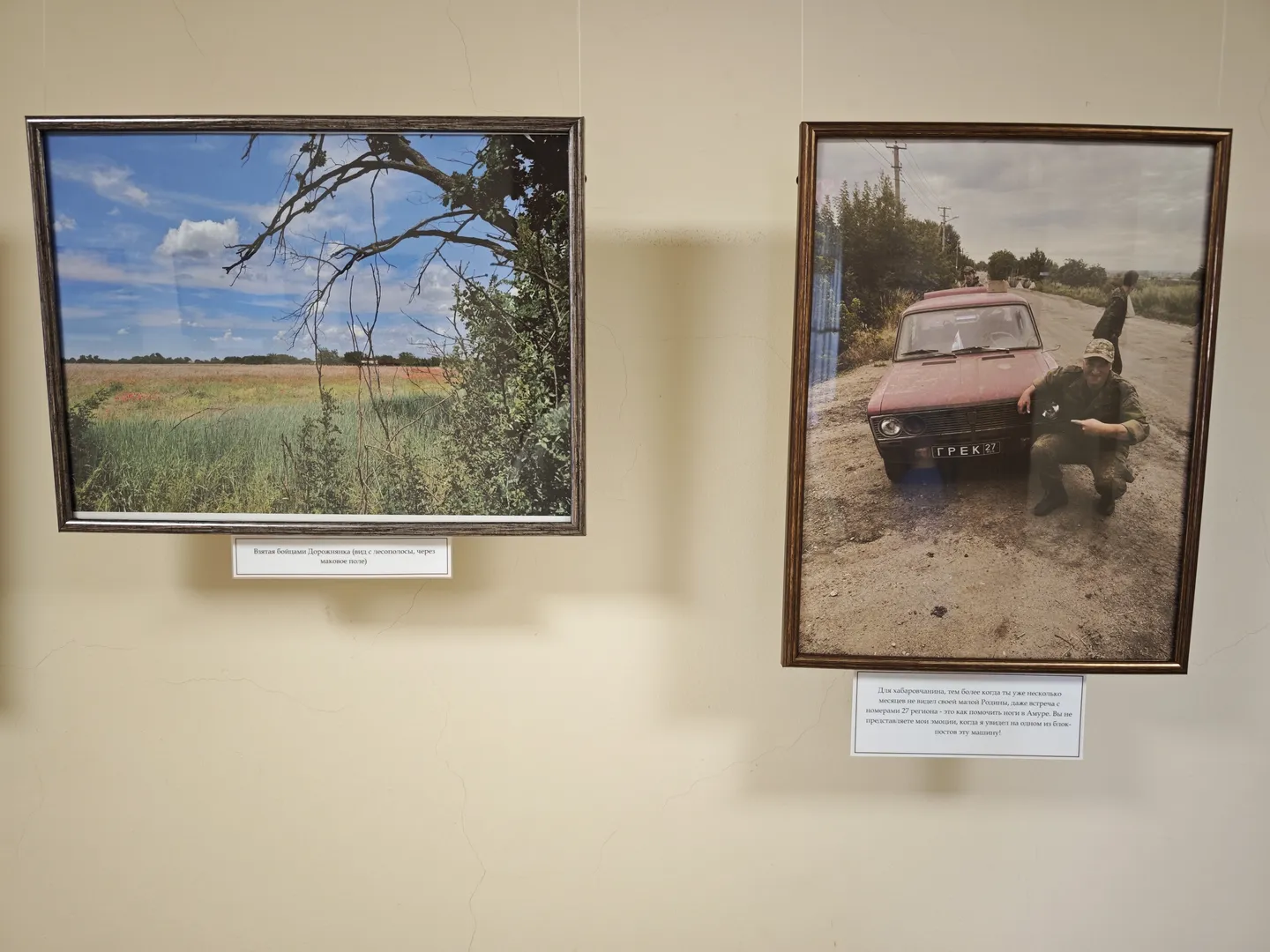 Two paintings on the wall. On the left, a field with trees; on the right, a person crouching beside a red car. Each painting has a white label with a description below.