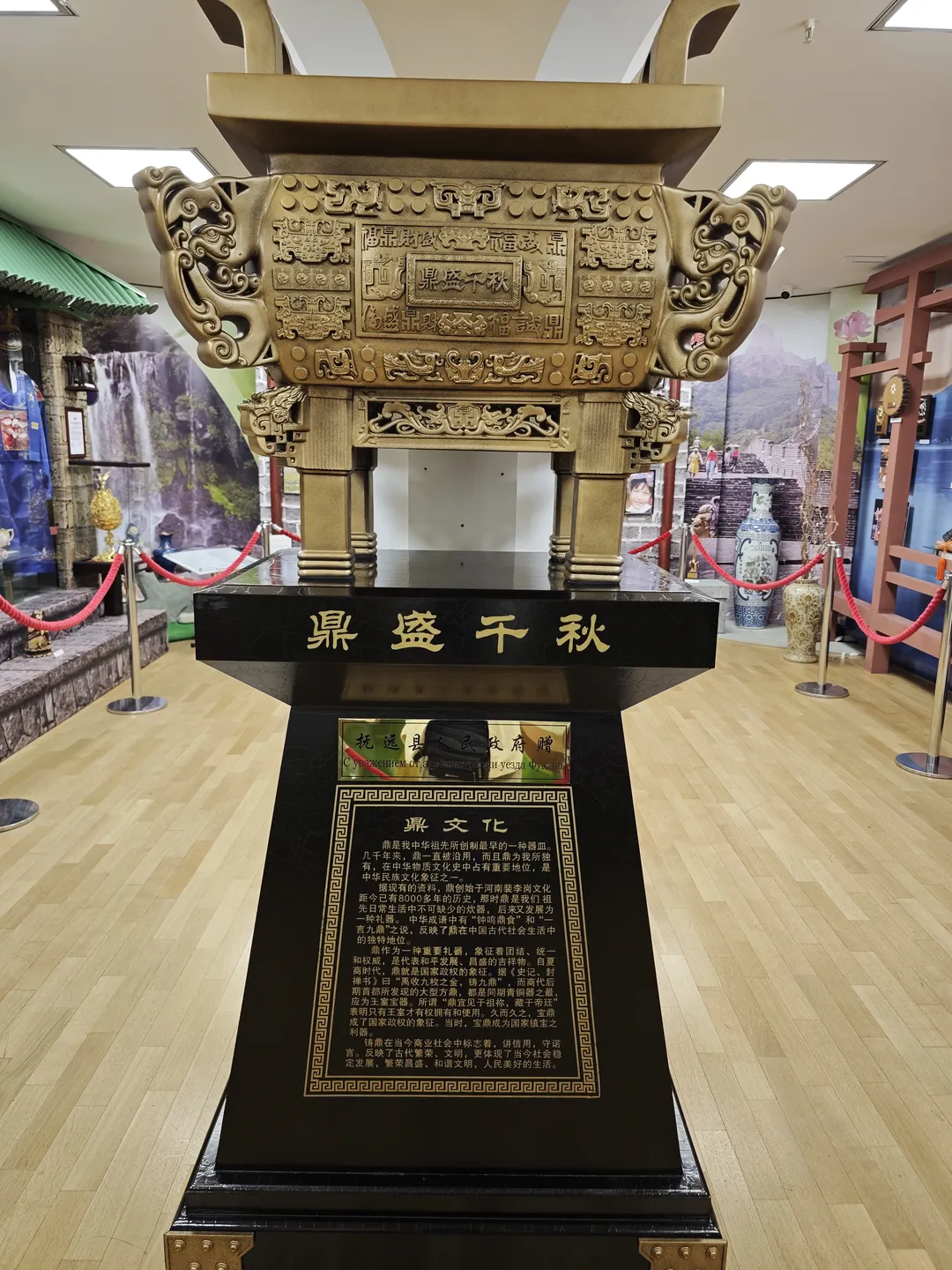 A large bronze tripod is placed on a black base, with golden text labels. The tripod is intricately carved with patterns, surrounded by a red rope barrier. The background features an indoor exhibition setting.