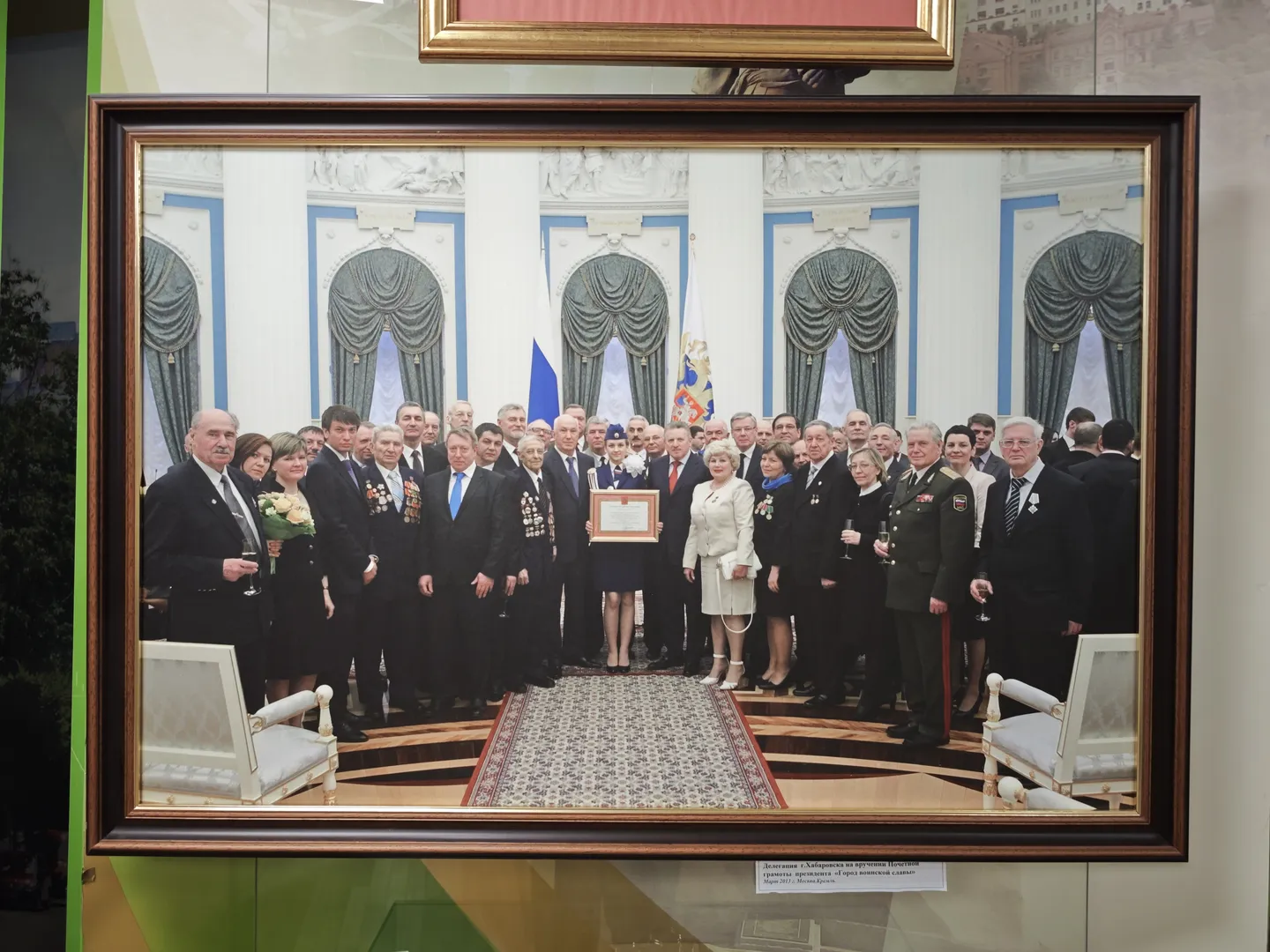 A group of people in formal attire posing for a photo indoors. The background features ornate windows and curtains. The person in the center holds a framed certificate, while others hold wine glasses. The photo is framed in a dark wood frame.