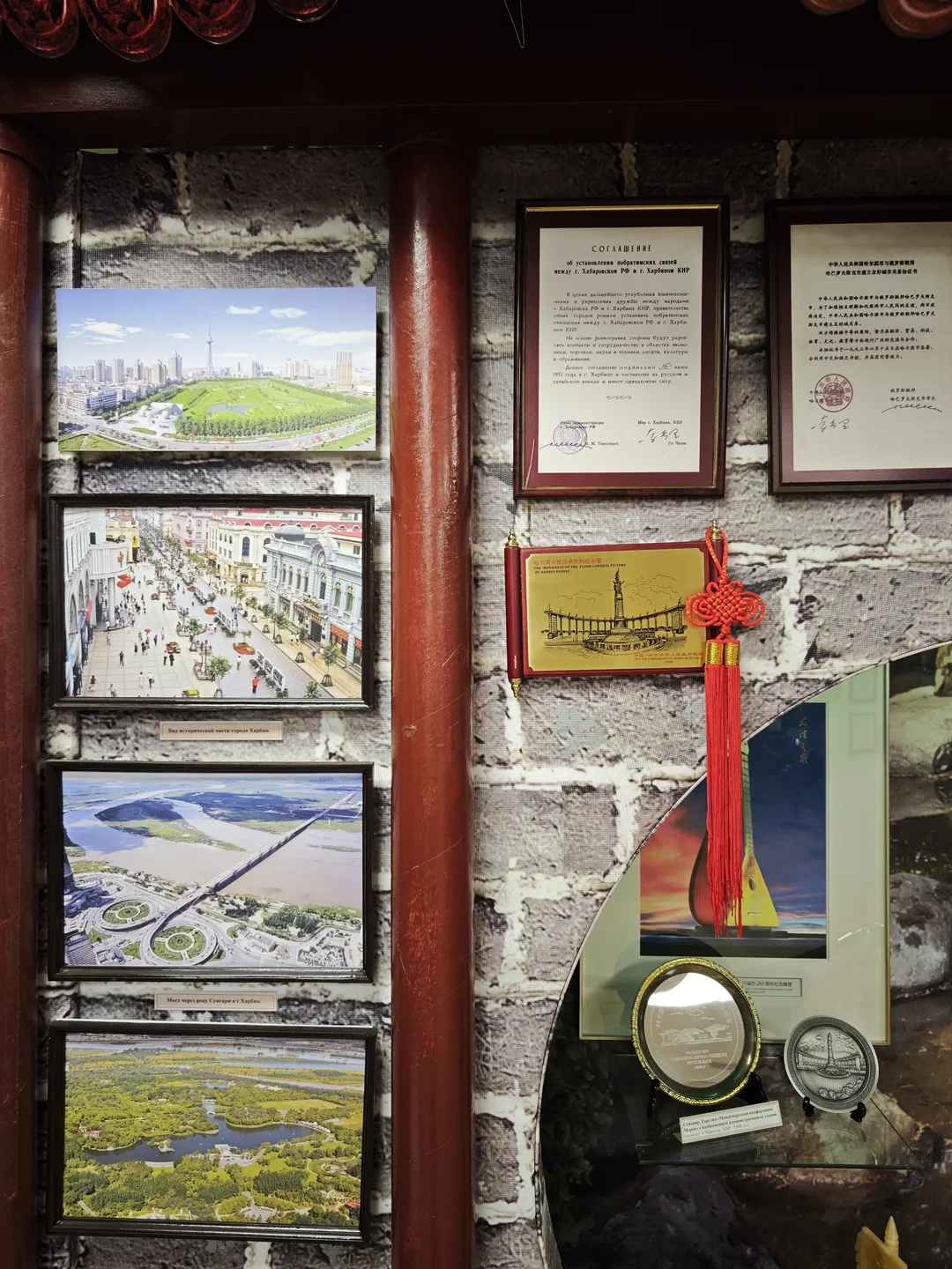 The wall features several cityscape photos and documents, with a red Chinese knot ornament hanging beside them. Below the photos, medals and souvenirs are displayed, and the wall tiles are gray. On the right side, there is a picture depicting musical instruments.