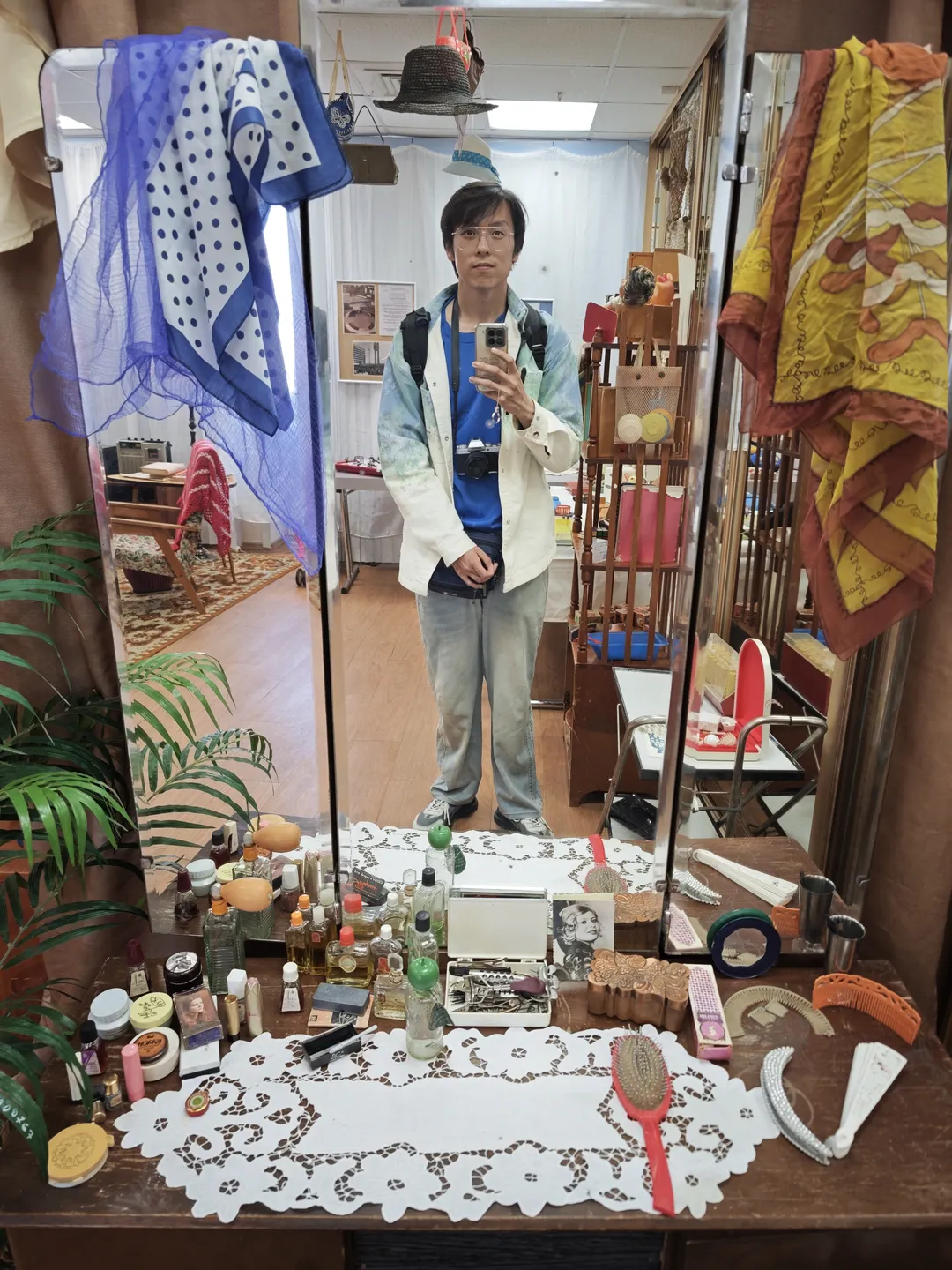 A person is standing in front of a mirror taking a selfie. Several scarves hang above the mirror. On the table beneath, various cosmetics and grooming items are neatly arranged. In the background, some furniture and decorations are visible.