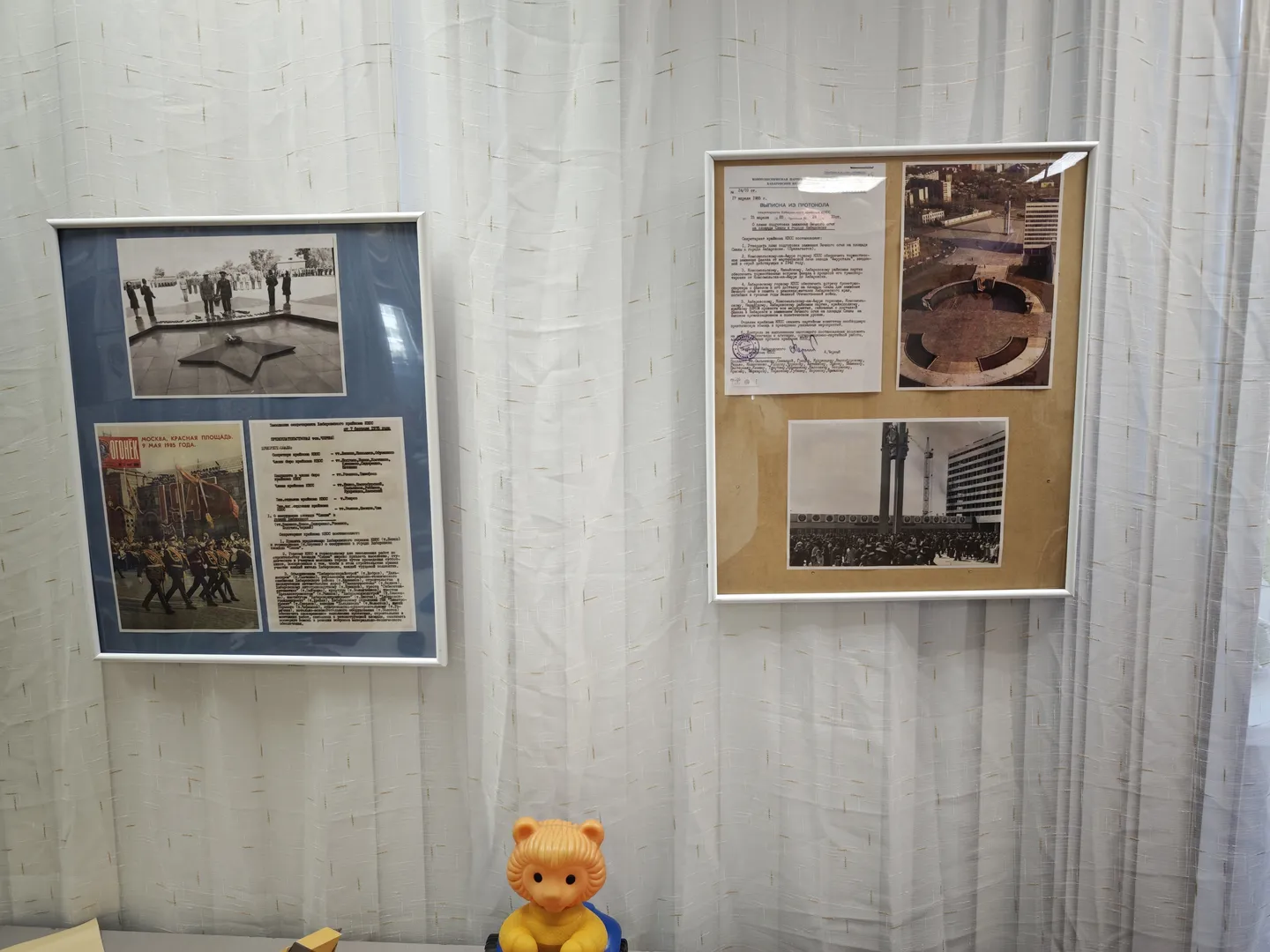 Two framed photos and documents are hung on the wall, depicting crowds and buildings. In the foreground, a yellow toy bear sits atop a blue object. The background features white curtains.