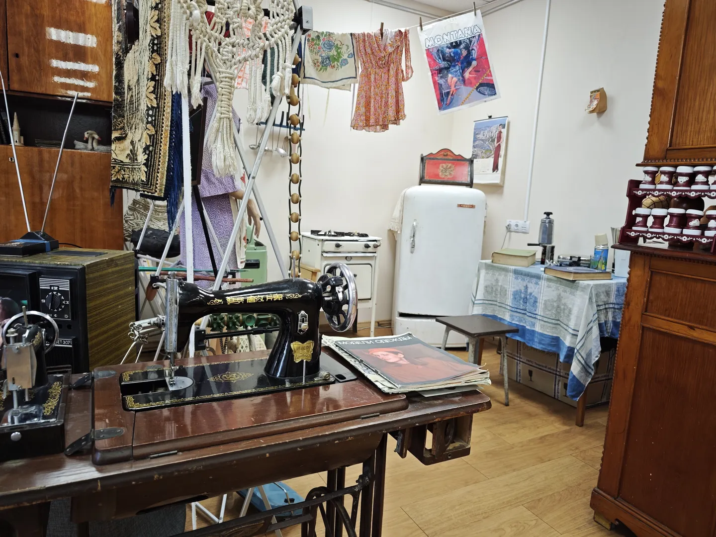 In the room, an old-fashioned sewing machine is placed on a wooden table with magazines piled beside it. In the background, a vintage refrigerator and a table covered with a blue-and-white checkered tablecloth are visible. Clothing and posters hang on the wall.