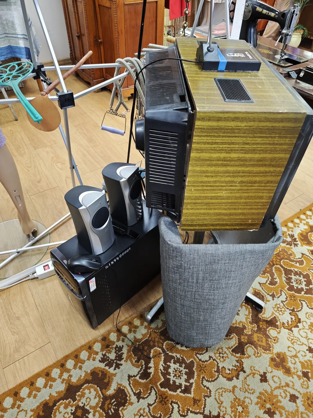 An old-fashioned television placed on a stand covered with a gray cloth. Multiple speakers are connected behind the TV, and below, a black equipment box holds a mouse and other items. The floor is covered with a patterned carpet.