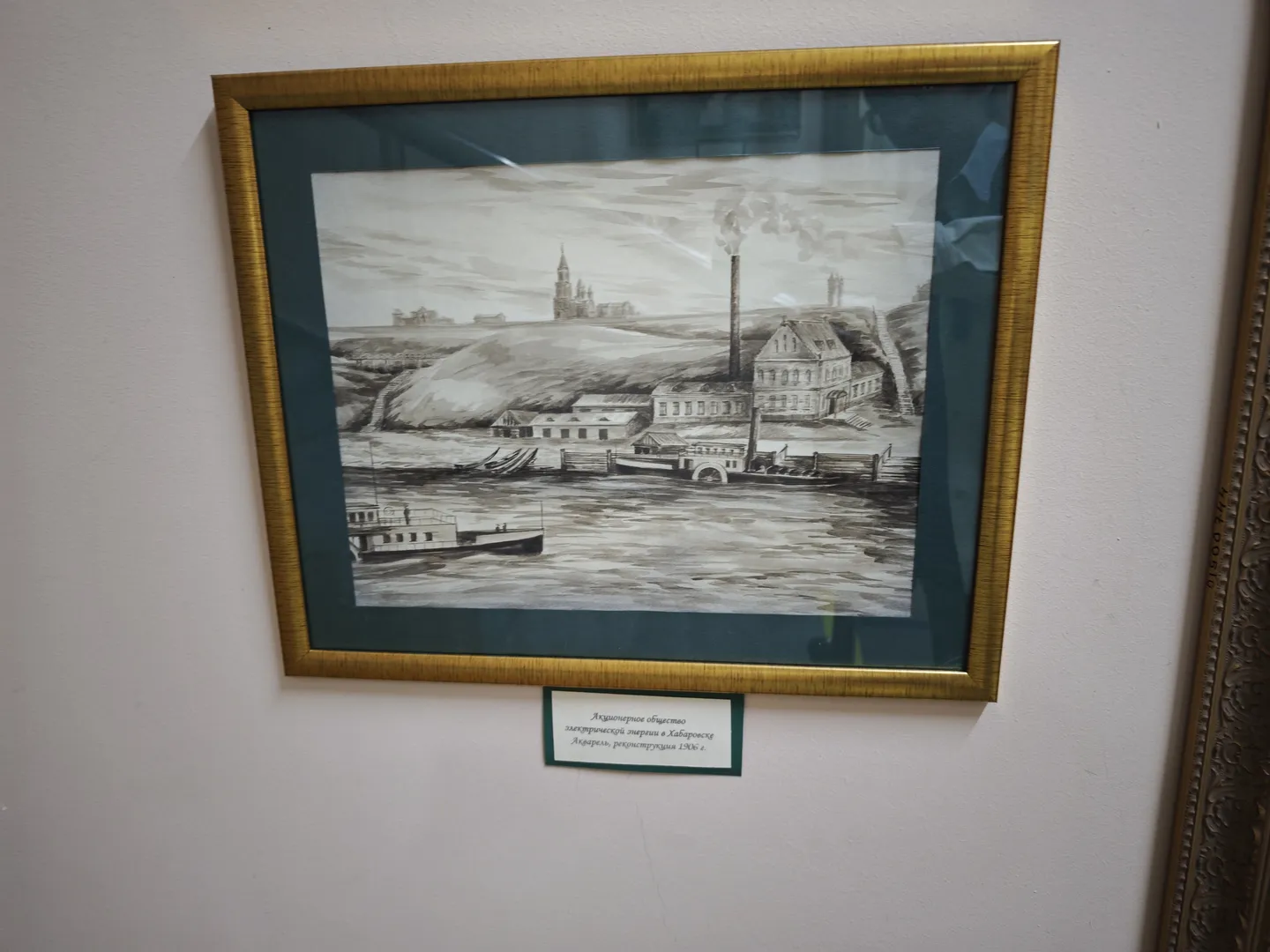 A black-and-white painting depicting a factory building by a river, with smoke rising from the chimney and boats on the river. The painting is framed in gold, with a label below. The background is a white wall.