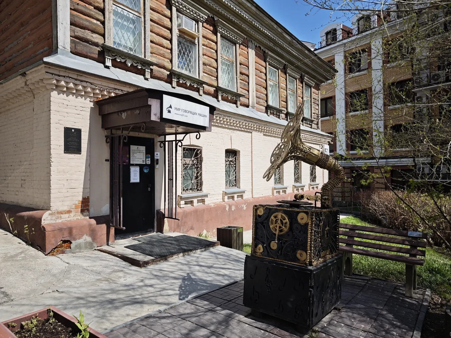 A wooden building with a black door at the entrance. Above the door, there is a sign hanging. In front, a metal sculpture resembling a bird with outstretched wings stands. Nearby, a long bench and some plants are visible.