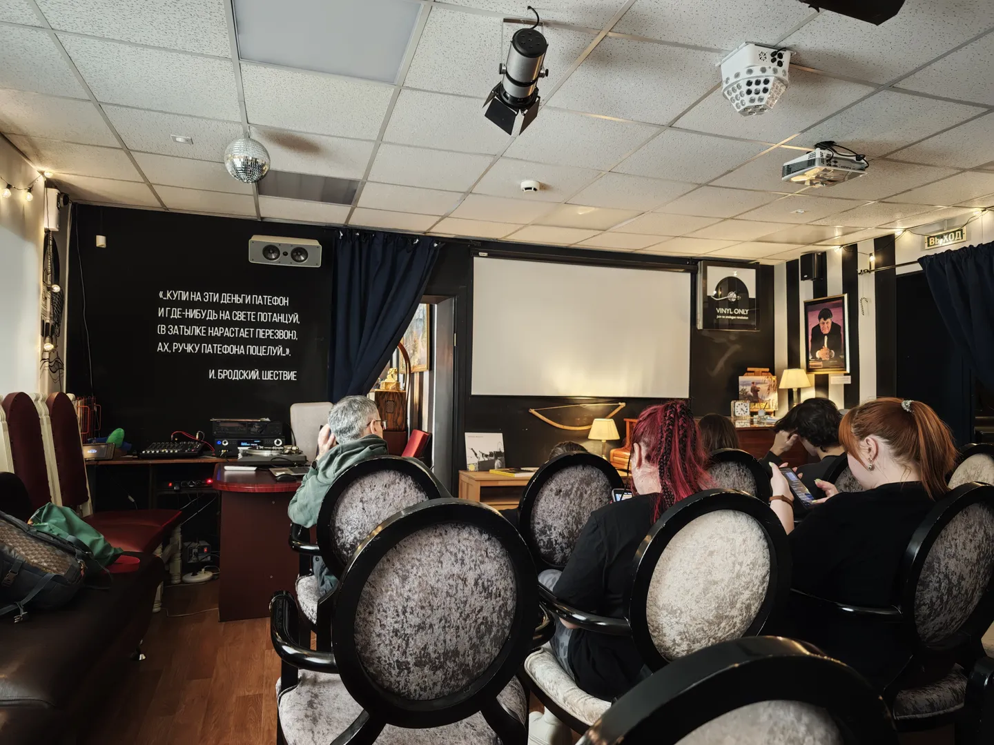 A room with several people seated on vintage chairs, facing a projection screen and sound equipment. Black curtains with Russian text hang on the wall, and various electronic devices are placed nearby. The ceiling features lighting and a projector.