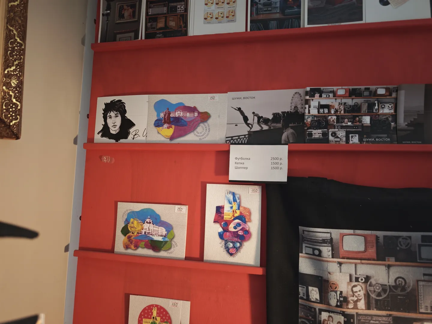 A red shelf displaying various items, including patterned cards and price tags. Above the shelf, black-and-white photos and colorful illustrations are shown. The background wall is light-colored.