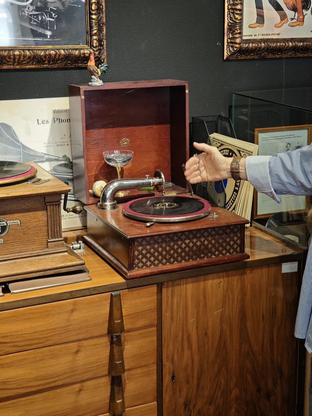 A wooden turntable is placed on a cabinet, with a hand operating it. The turntable has a vintage design, complete with a metal horn. Other similar devices and decorative items are visible in the background.