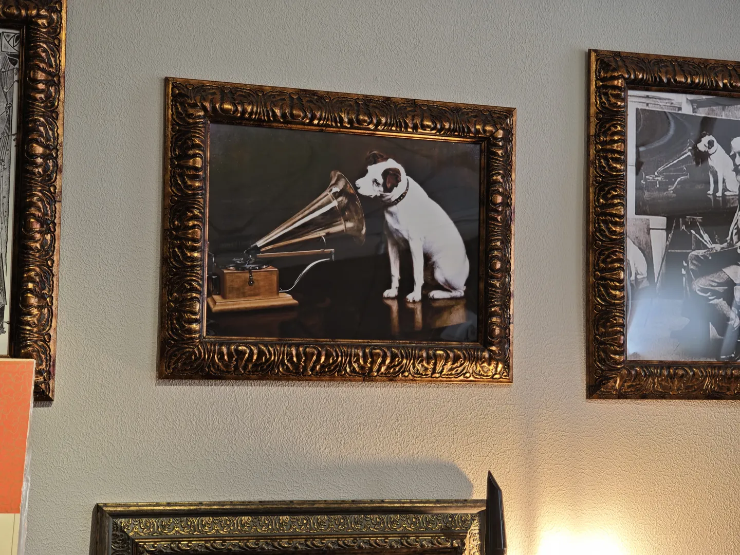 A painting in a golden frame on the wall depicts a dog facing a turntable. On either side of the painting, other similar artworks are displayed. The background wall is light-colored.