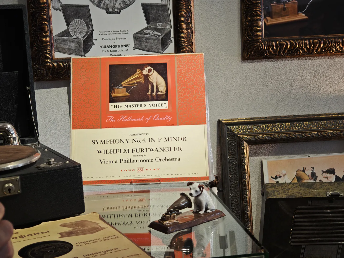 A record album cover displayed in front of a glass cabinet, featuring a dog facing a turntable. Inside the cabinet, there are small turntable models and other items. Related posters are hung on the background wall.