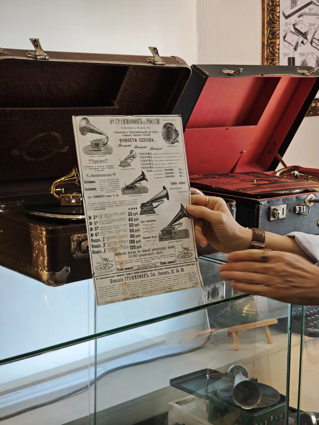 A hand holding a piece of paper featuring various turntable designs, displayed in front of a glass cabinet. The cabinet contains different turntable models. In the background, an open suitcase is visible.